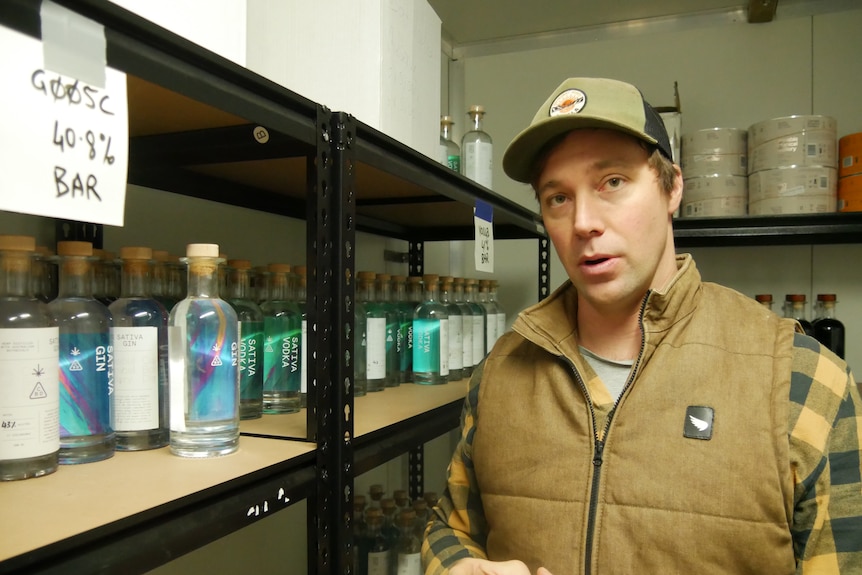 a man wearing a cap, checked short and vest stands in a room with bottles of sativa gin
