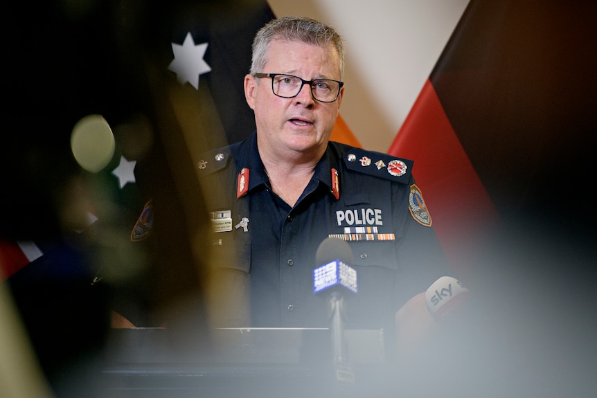 A man wearing a navy police uniforms talks to microphones. He has grey hair and black glasses.