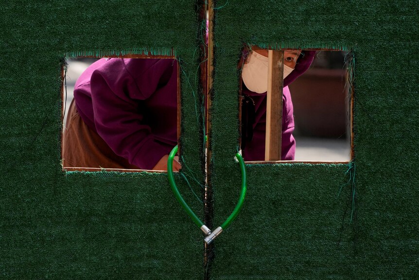 A woman looks through cutouts in a door locked with a bike lock.