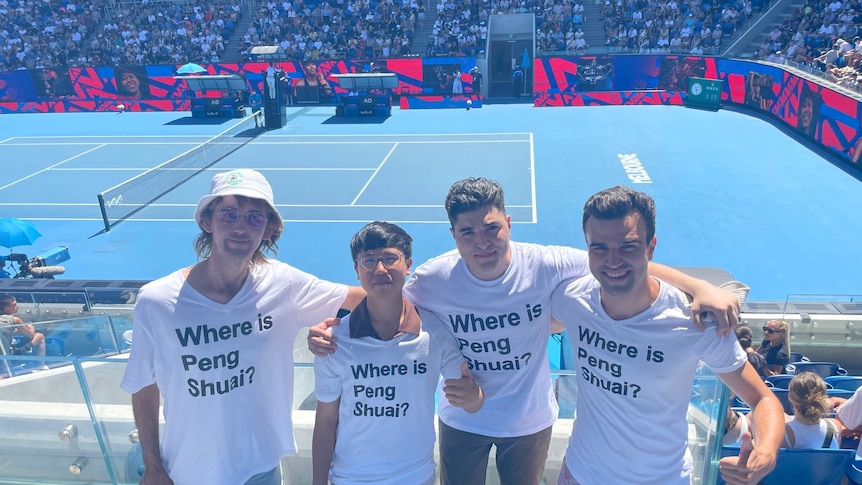 Four 30-something men wearing 'Where is Peng Shuai' T-shirts link arms with Australian Open tennis court behind them
