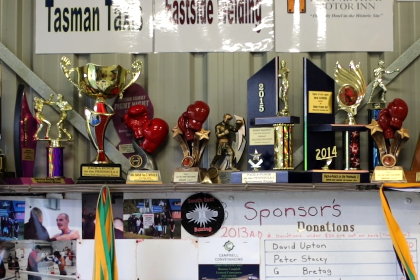 A range of boxing trophies stand on a shelf inside the gym along a wall as photos surround them.