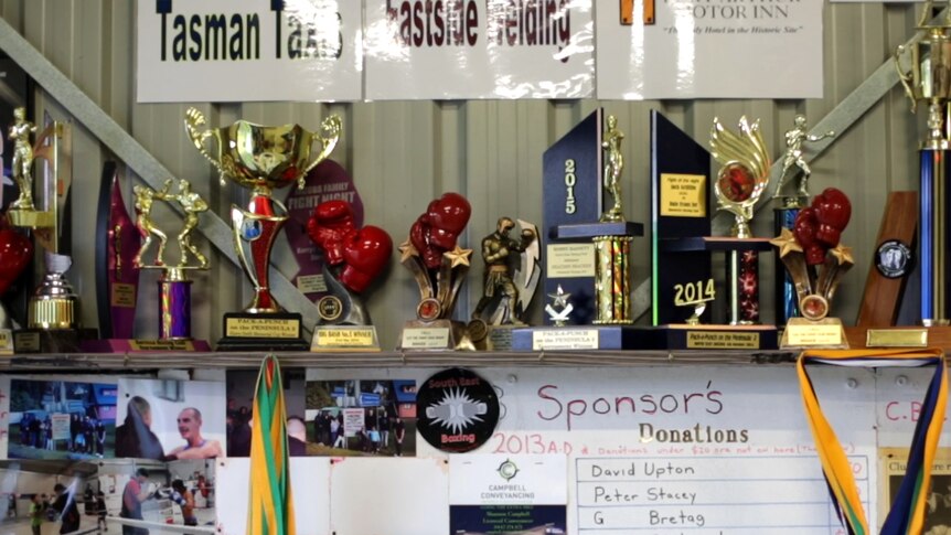 A range of boxing trophies stand on a shelf inside the gym along a wall as photos surround them.