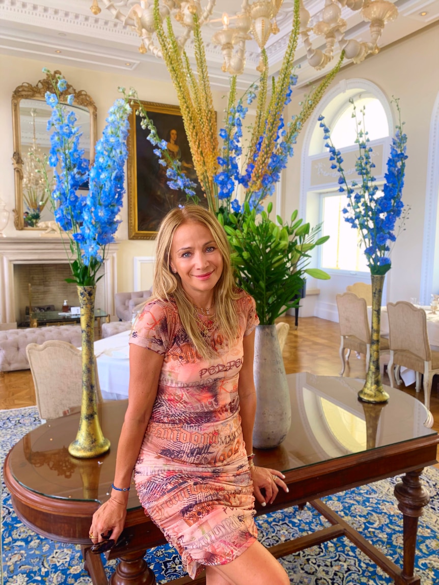 A middle-aged woman with sandy blonde hair leans against a table in a beautifully decorated room with blue flowers. 