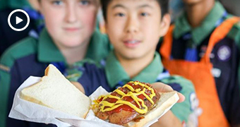 Two young boys, blurred in background, hold forward a sausage sizzle sandwich covered in sauce, clear and in the foreground.