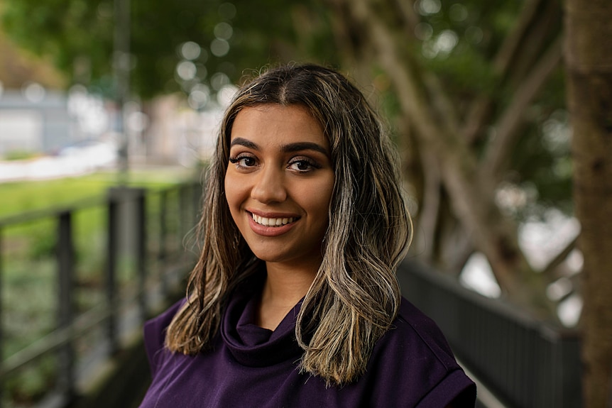 Young Muslim woman Sabreen Hussain smiling, wearing purple top, with trees in background.