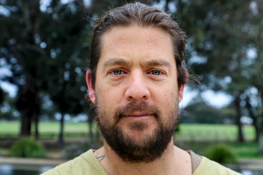 Man stares down camera with trees in background.