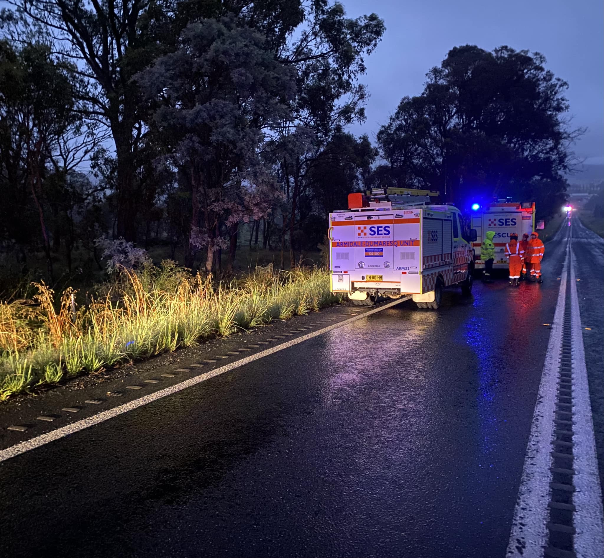 Bright red and blue lights flash on a wet roadsdie where two SES trucks are parked with emergency workers on the road