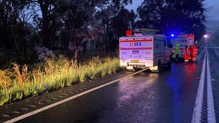 Bright red and blue lights flash on a wet roadsdie where two SES trucks are parked with emergency workers on the road