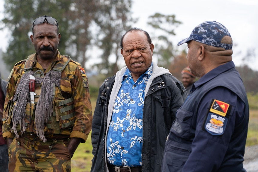 Police officer speaks to a local man in a field, with defence personnel next to him.