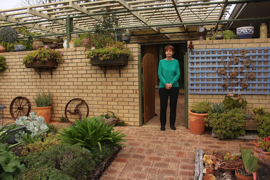 Sue stands near a gate out the front of her house, in her garden with lots of potted plants