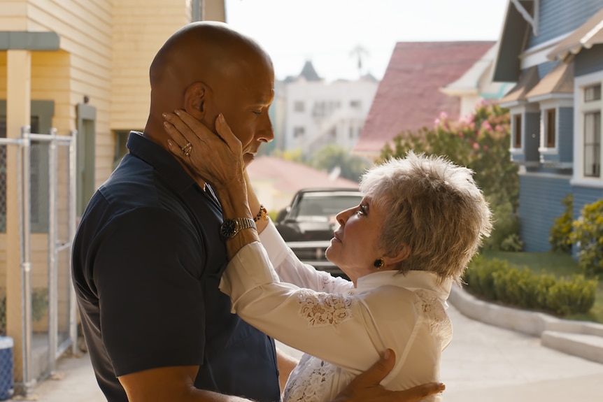 A bald black man in a navy polo stands with a shorter elderly white woman with grey hair who is cupping his face.
