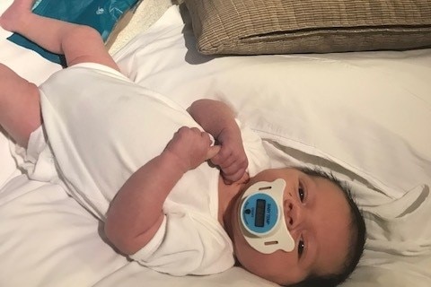 A small baby in a white onesie lays on a bed with a dummy in her mouth