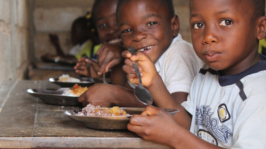 Three young school boys in Haiti look at the camera holding spoons and bowls of food in their hands.