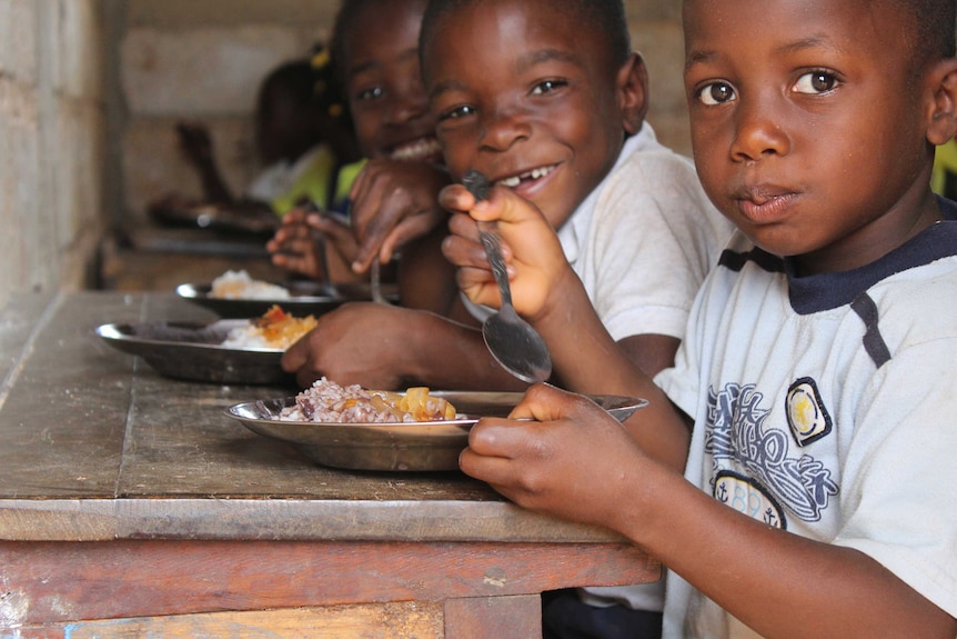 Three young school boys in Haiti look at the camera holding spoons and bowls of food in their hands.