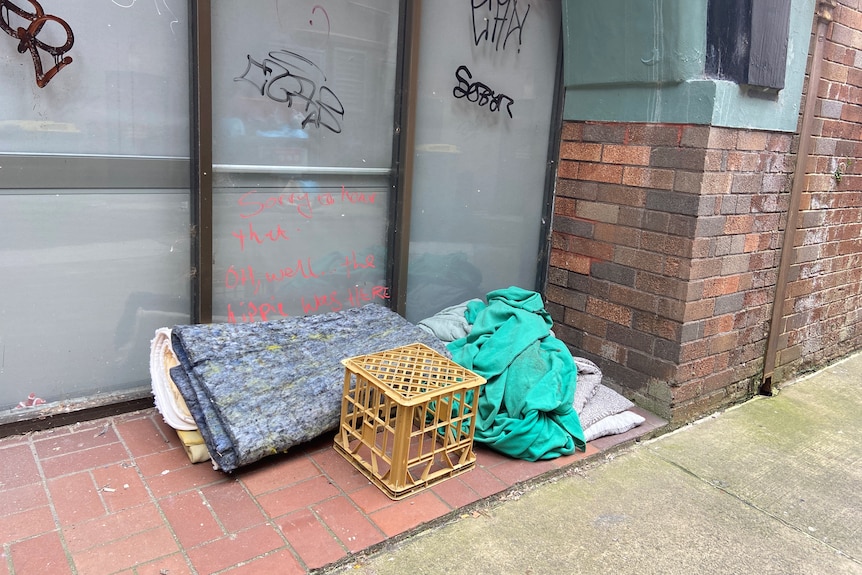A milk crate and bedding in lying on the street in front of a disused shop.