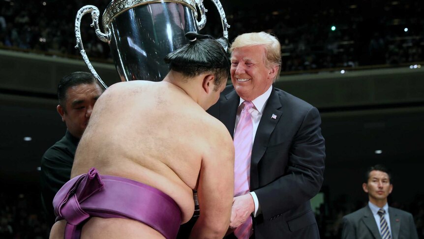 Trump smiles as he hands over giant silver trophy to Japanese sumo wrestler.