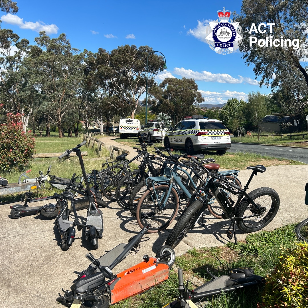 A pile of bikes lined up with a scooter on the ground, and police cars in the background