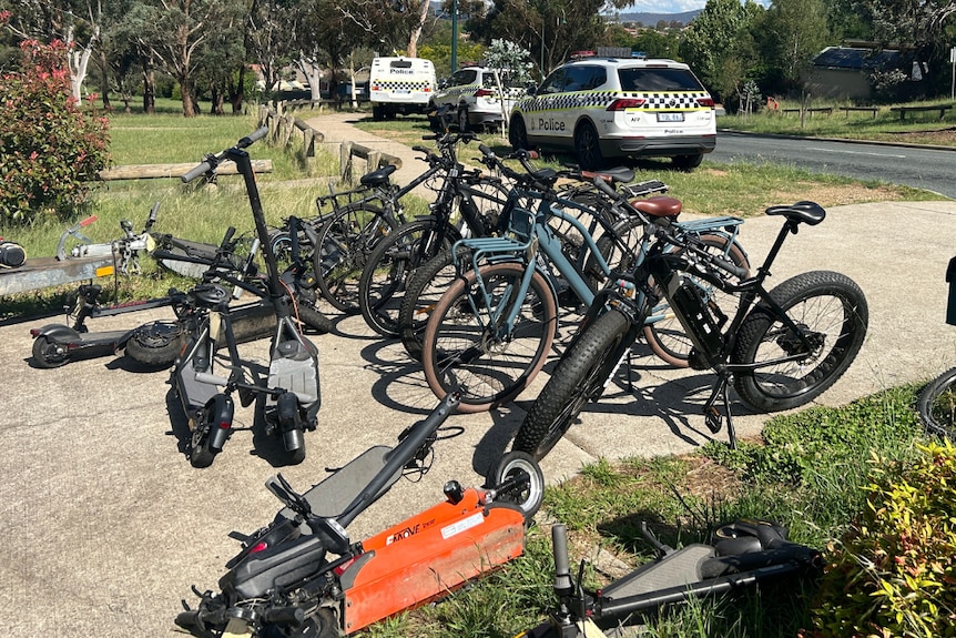 A pile of bikes lined up with a scooter on the ground, and police cars in the background