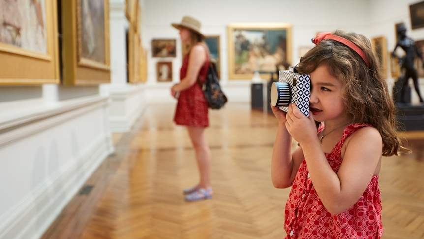 A young girl in a room of paintings takes a photo with a camera.