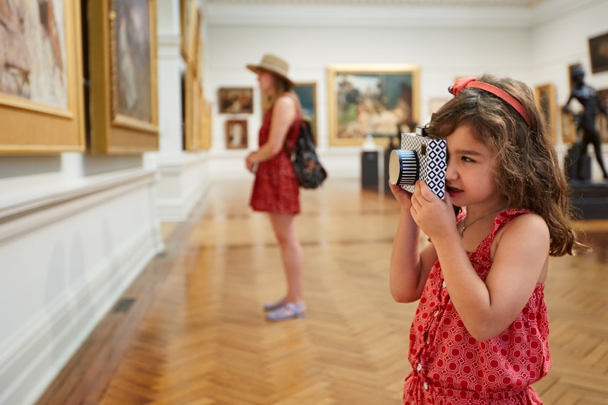 A young girl in a room of paintings takes a photo with a camera.