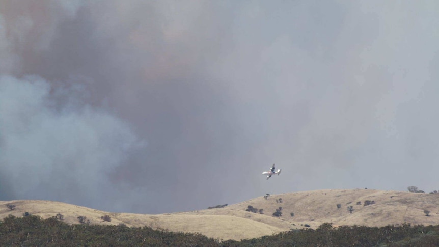 A firefighting plane flying over hills with smoke in the background.