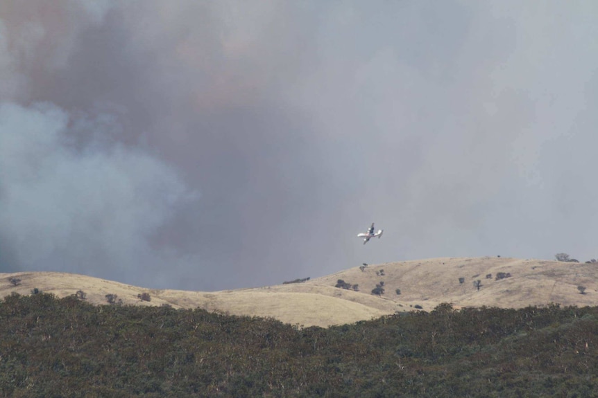 A firefighting plane flying over hills with smoke in the background.