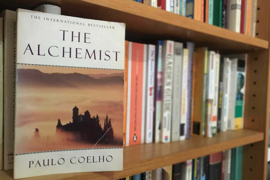 Close up of The Alchemist book cover with blurred books on a shelf behind it.