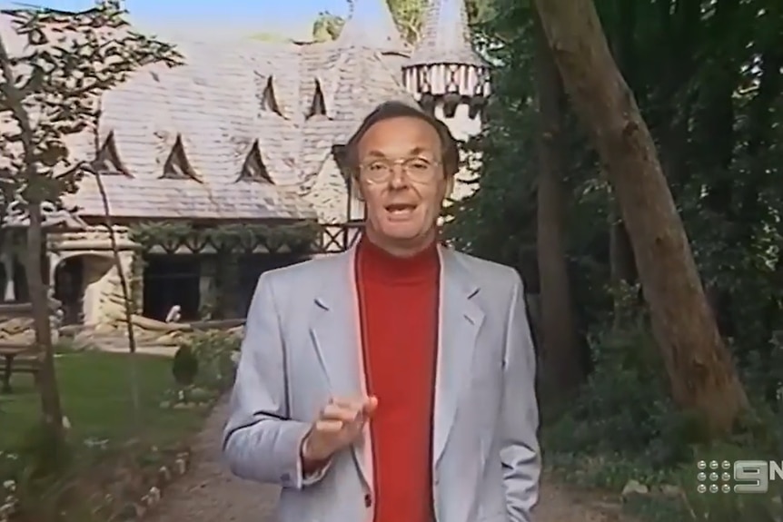 Jeff Watson wears a dark red skivvy with a grey jacket while standing in what appears to be a tropical resort
