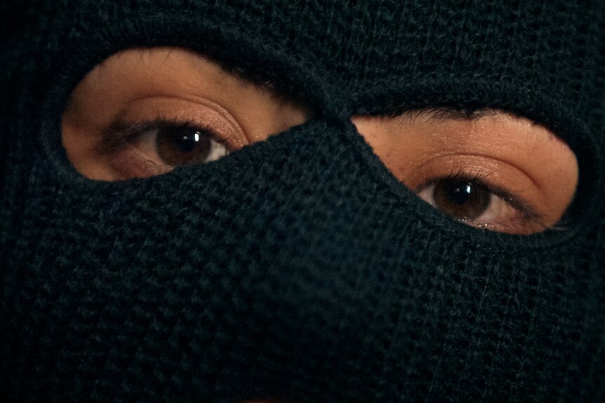A woman's eyes look at the camera. She is wearing a black balaclava and the rest of her face cannot be seen.