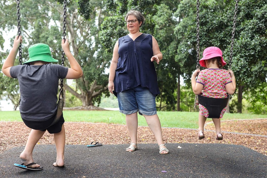 Brisbane mother Jenny Griffin stands in front of her children, 6 and 8, who are on swings at a park.
