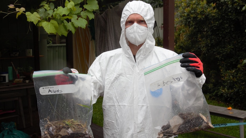 A man in a white hazmat suit with mask holding two sandwich bags labelled 'asbestos'