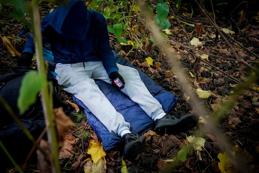 A young man in a hoodie sits on his sleeping bag in a park while taking a can of tuna out of his backpack.