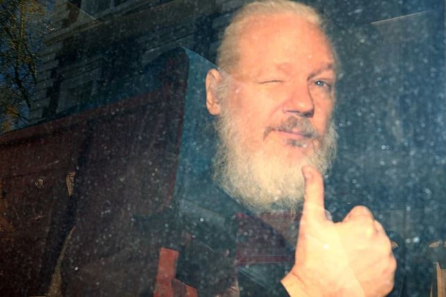 Julian Assange holding a thumbs up while looking out the window of a police van after his removal from the Ecuadorian embassy.