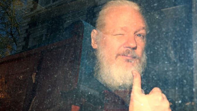 Julian Assange holding a thumbs up while looking out the window of a police van after his removal from the Ecuadorian embassy.