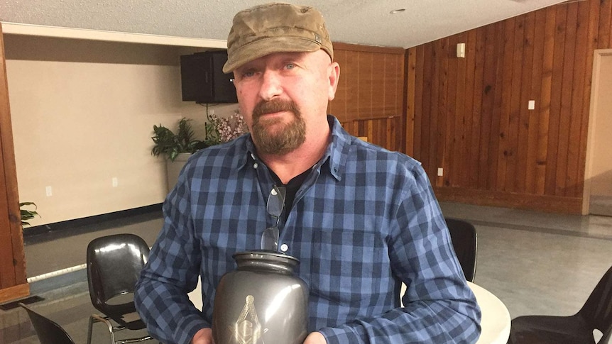 A man standing in a small auditorium holding an urn of ashes