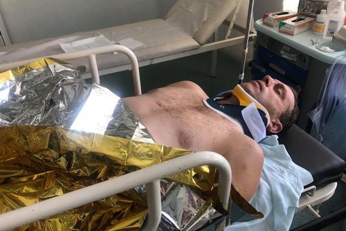 Man lies on hospital bed, with eyes closed and in a neck brace.