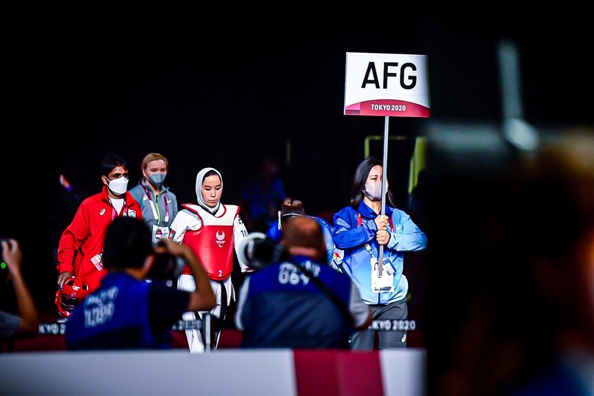 A small sports team headed by masked woman in blue carrying a flag that says 'AFG' marches at night.