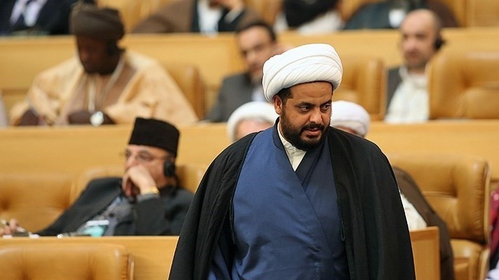 In a tan conference hall, Iraqi MP Qais Khazali wears a black cape and looks to the right of the frame.