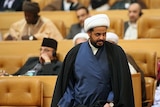 In a tan conference hall, Iraqi MP Qais Khazali wears a black cape and looks to the right of the frame.