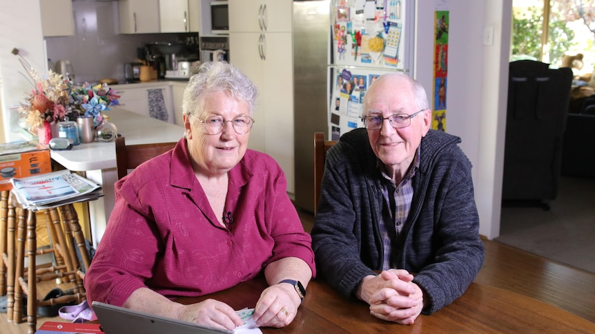 An older woman and an older man sit at dining table, both wearing warm jumpers and reading glasses