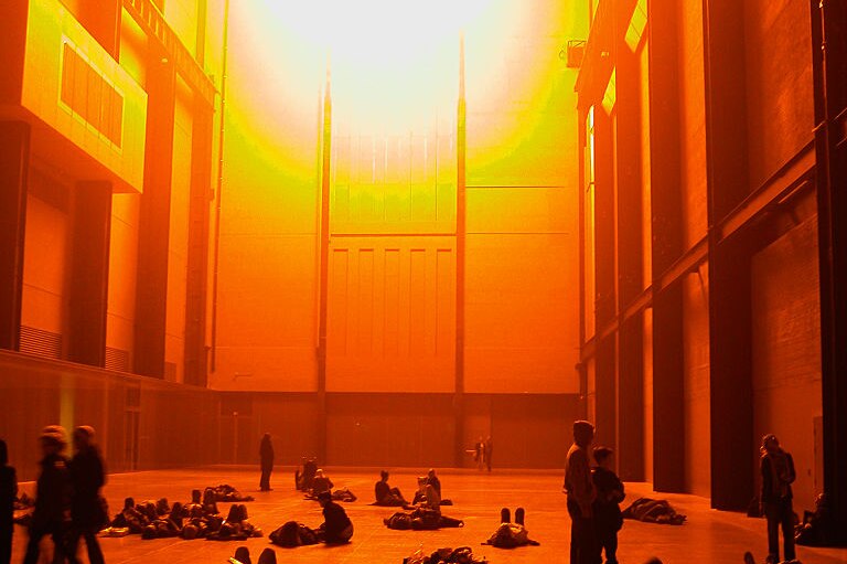 People inside a high-ceilinged room bathed in orange light shining in a big orb shape like the sun