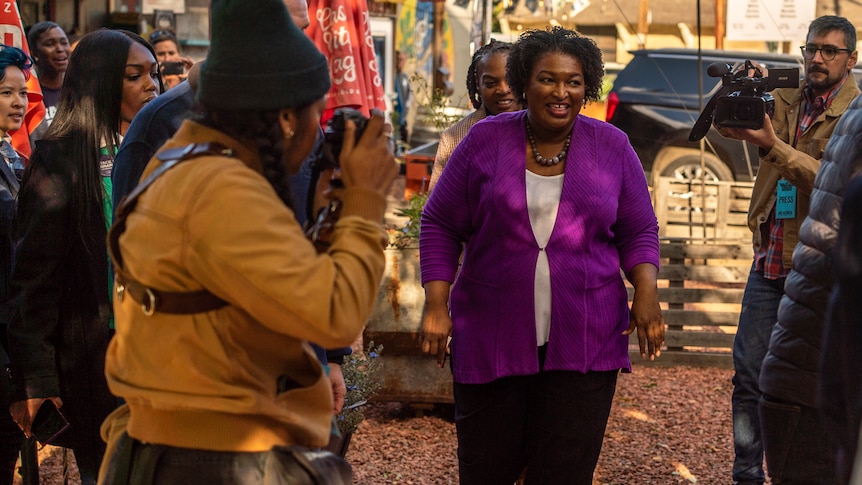 Stacey Abrams smiles while wearing a purple suit and walking through a group of photographers