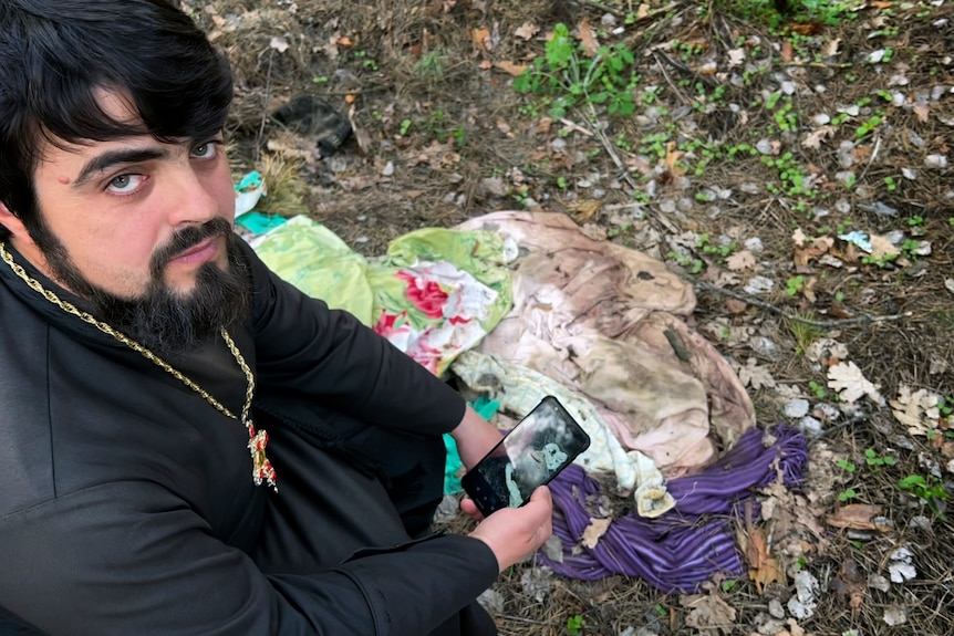 A bearded priest holds a phone by a pile of torn clothes 