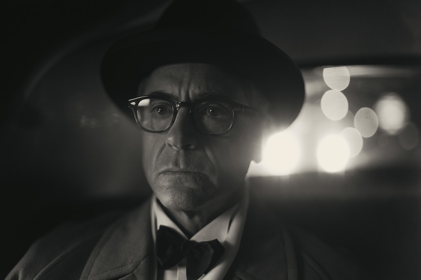 A black and white shot of a middle-aged white man with glasses sitting in a car wearing a suit with a hat and bow-tie.