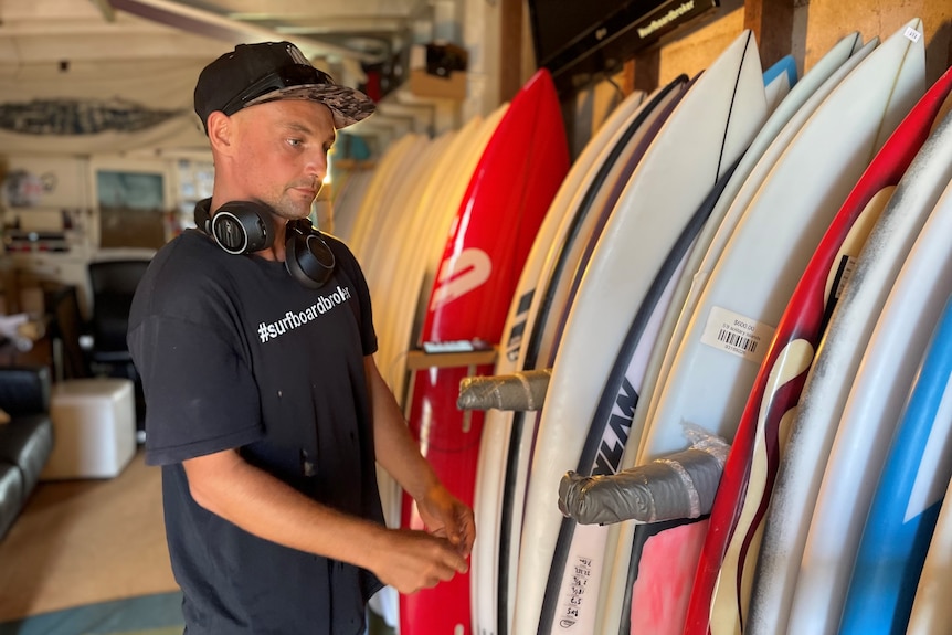 A man wearing a t-shirt and cap stands looking at a row of surfboards inside a shed.