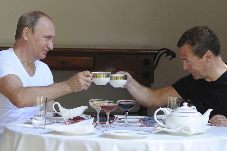 Vladimir Putin and Dmitry Medvedev cheers with tea cups while smiling at each other over a breakfast spread