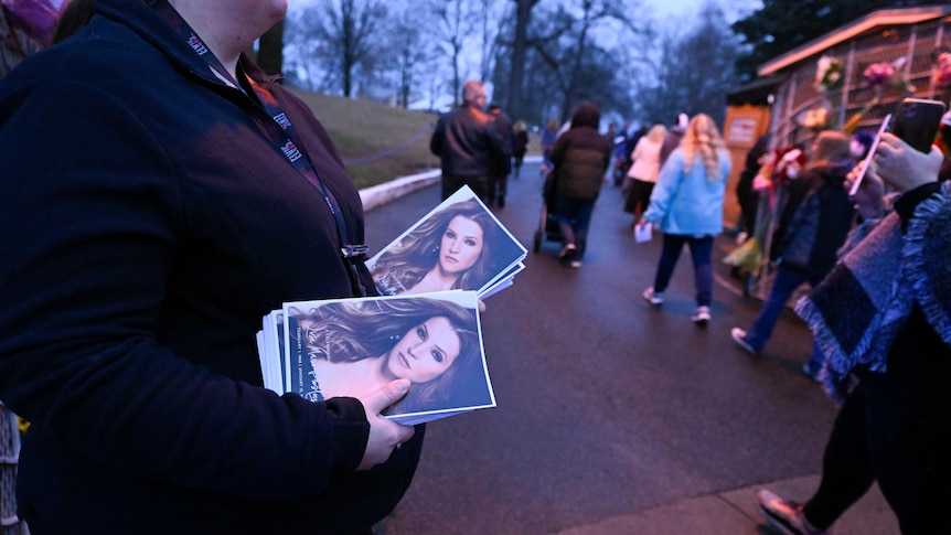 Attendant holds memorial service program with Lisa Marie Presley's photo, as people walk into Graceland.
