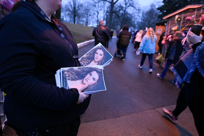 Attendant holds memorial service program with Lisa Marie Presley's photo, as people walk into Graceland.