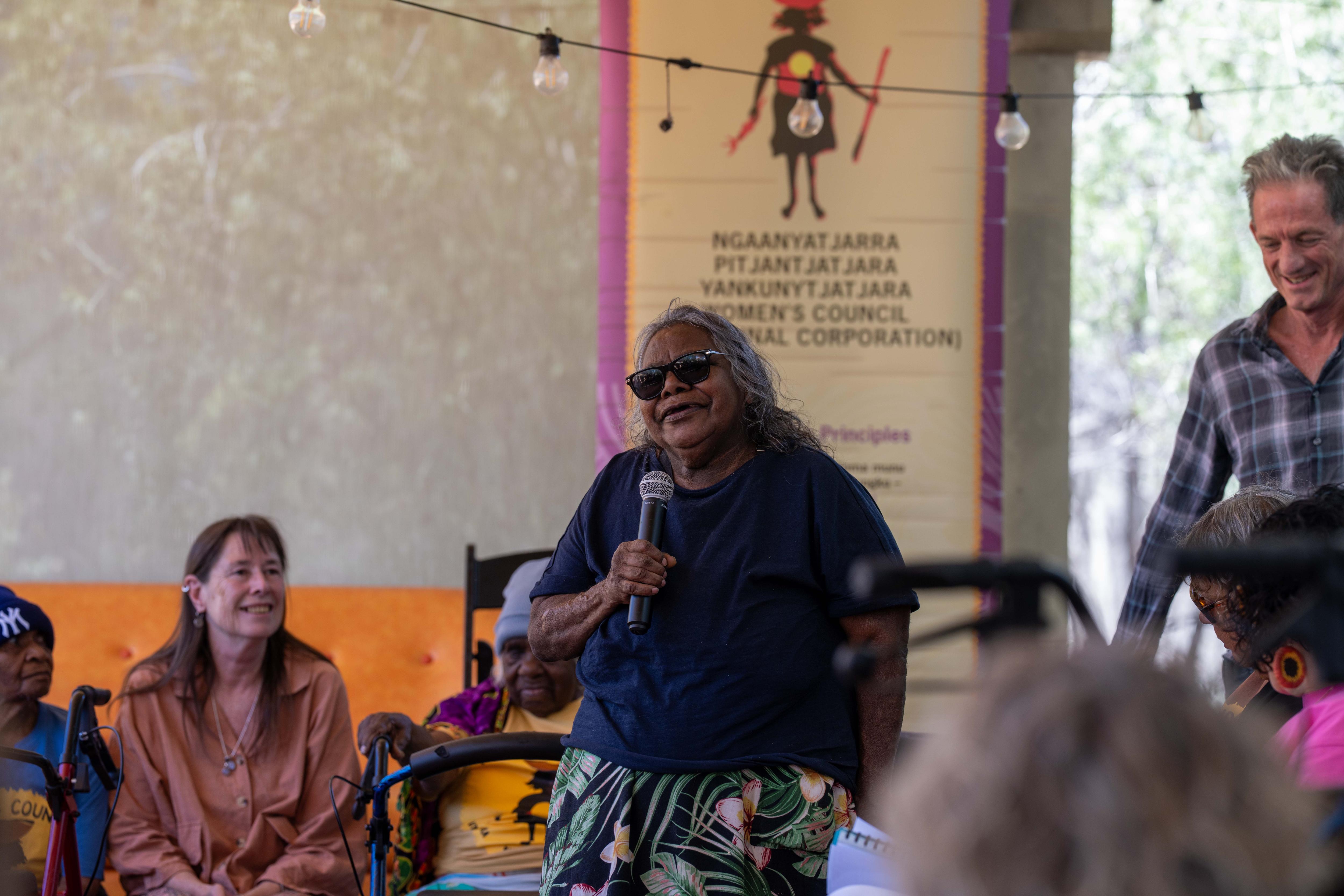 Una mujer aborigen con una camisa negra se encuentra entre la multitud y sostiene un micrófono.