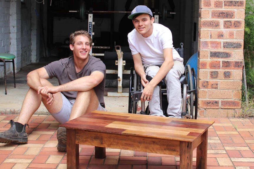 Two young men in front of a wooden coffee table. One man sits on the paved ground, the other leans forward in a wheelchair.